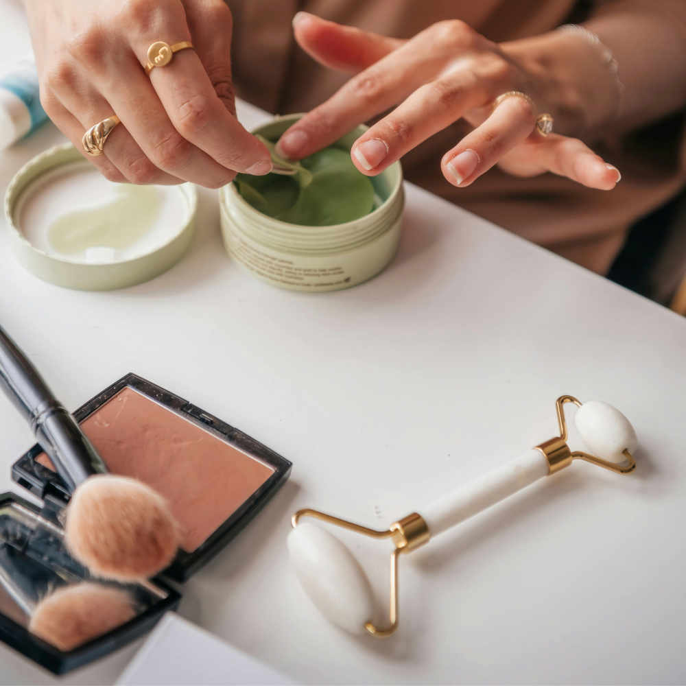 Person applying cream to a small container with makeup tools on a table.