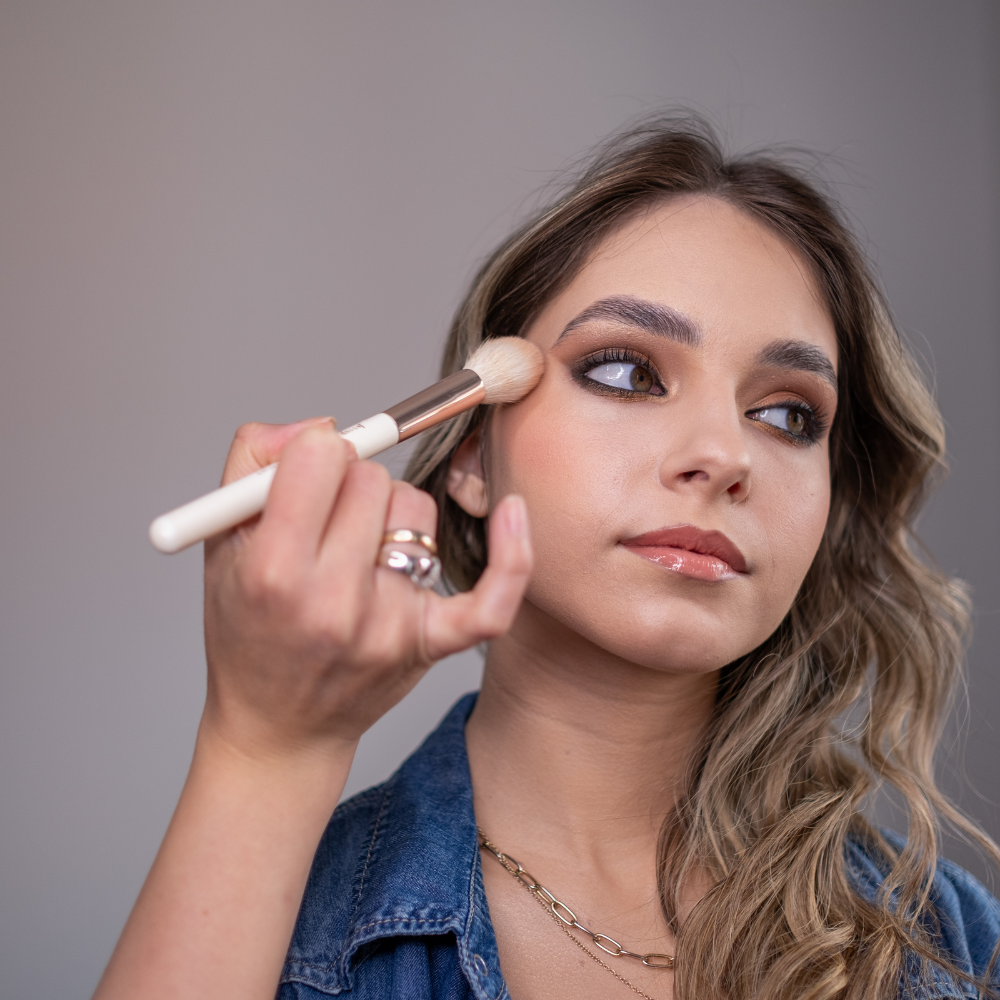 Woman applying makeup with a brush against a neutral background

